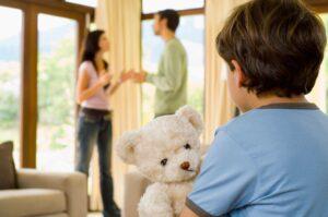 A young boy looks at his arguing parents while holding a soft teddy bear close to his chest